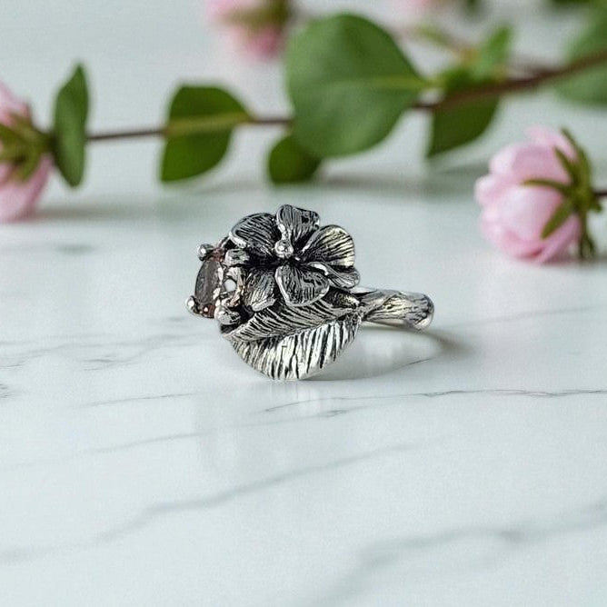 Silver floral ring on a marble surface with pink flowers in the background