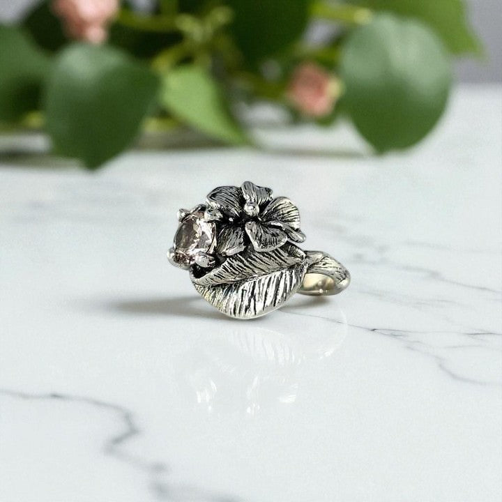 Silver ring with a central gemstone on a marble surface with green foliage in the background