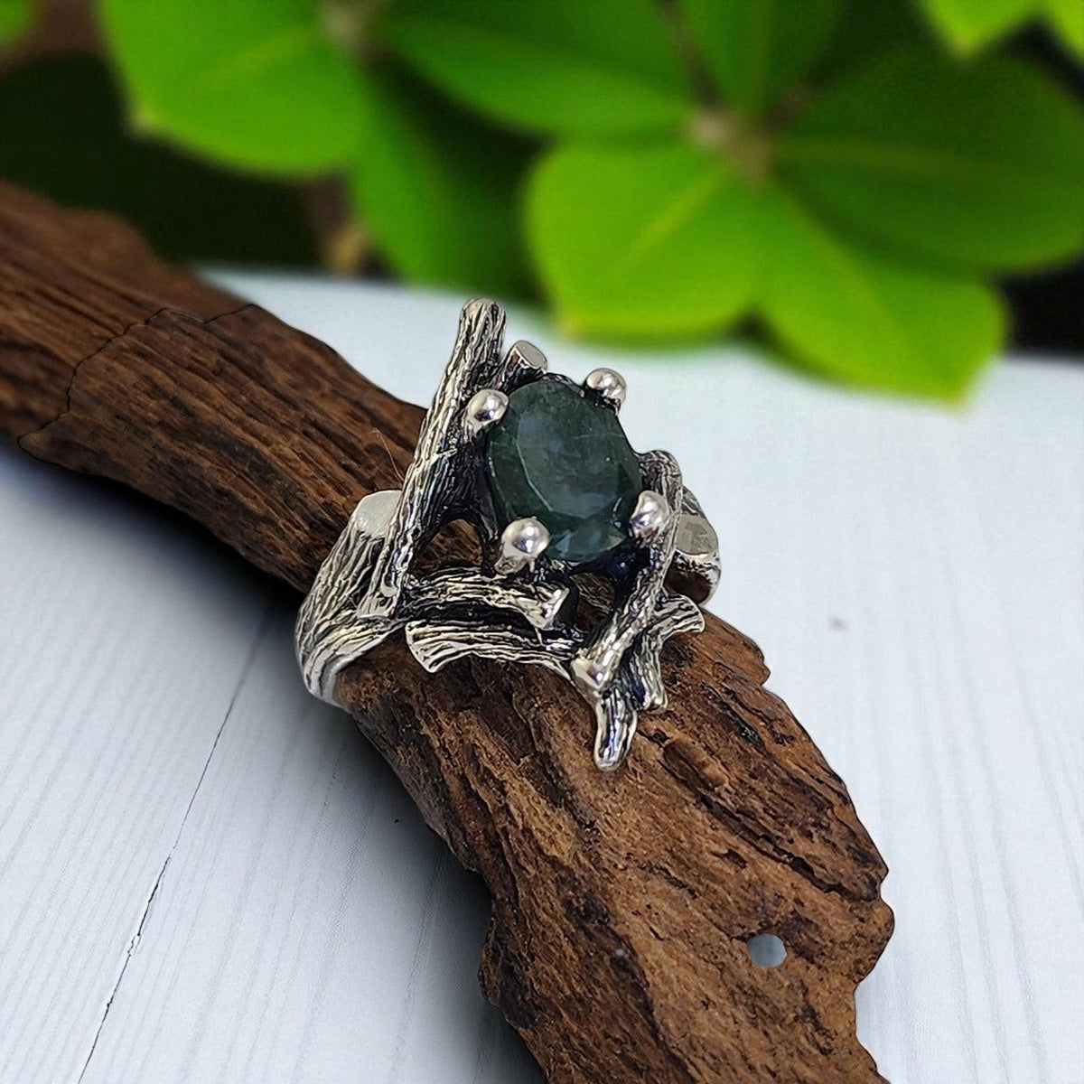 Silver ring with a dark gemstone on a wooden branch with green leaves in the background