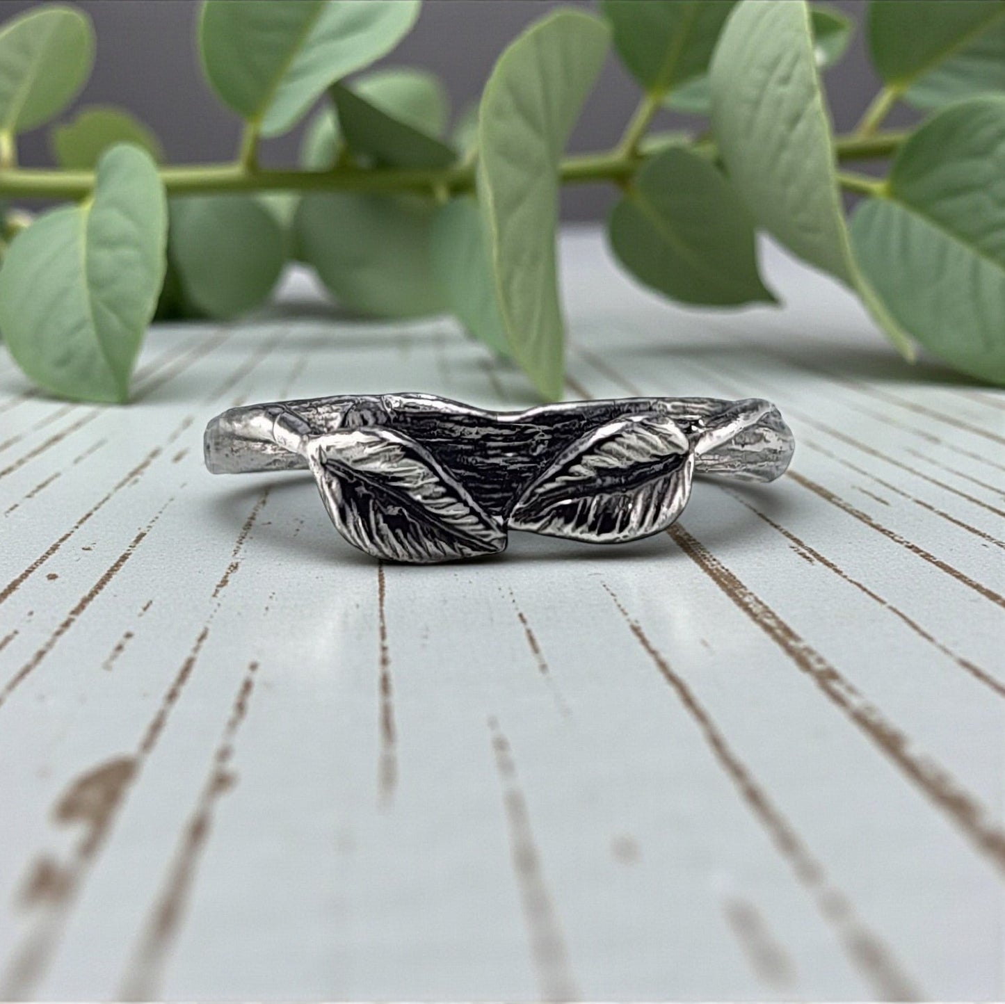 Silver leaf-shaped ring on a wooden surface with green leaves in the background