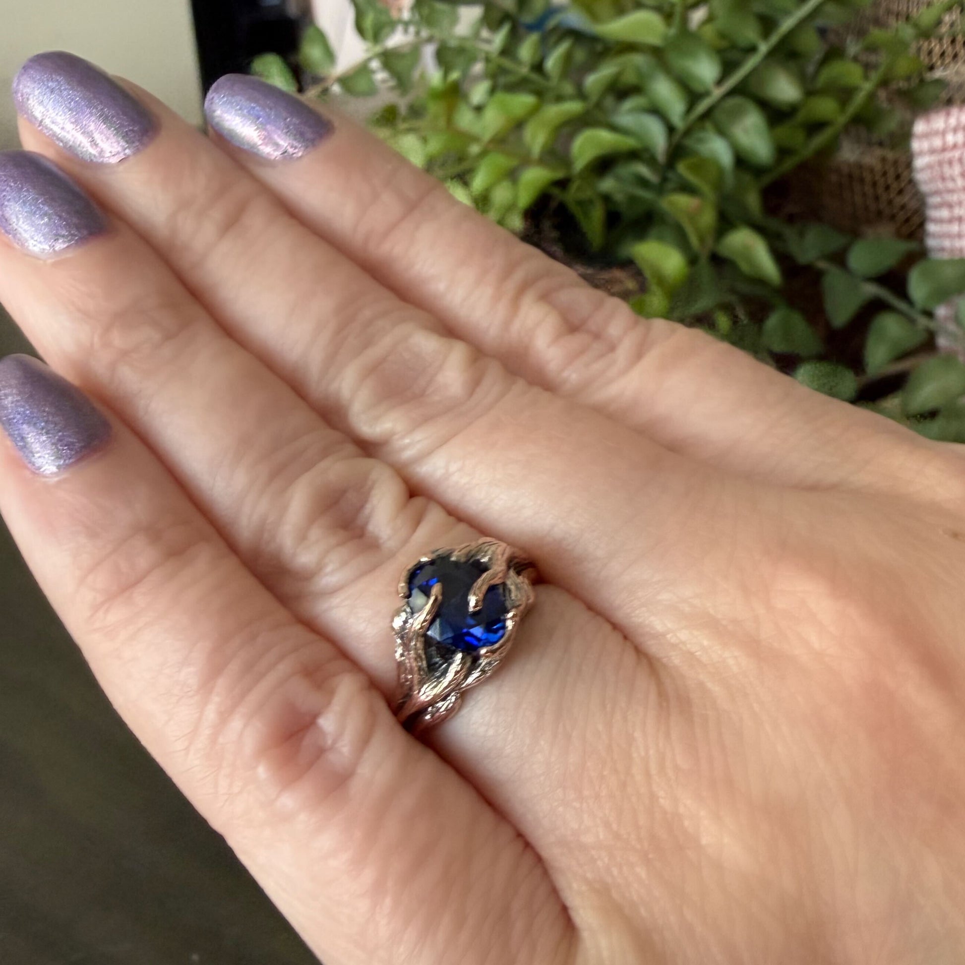 Hand wearing a ring with a blue gemstone, blurred green plant in the background