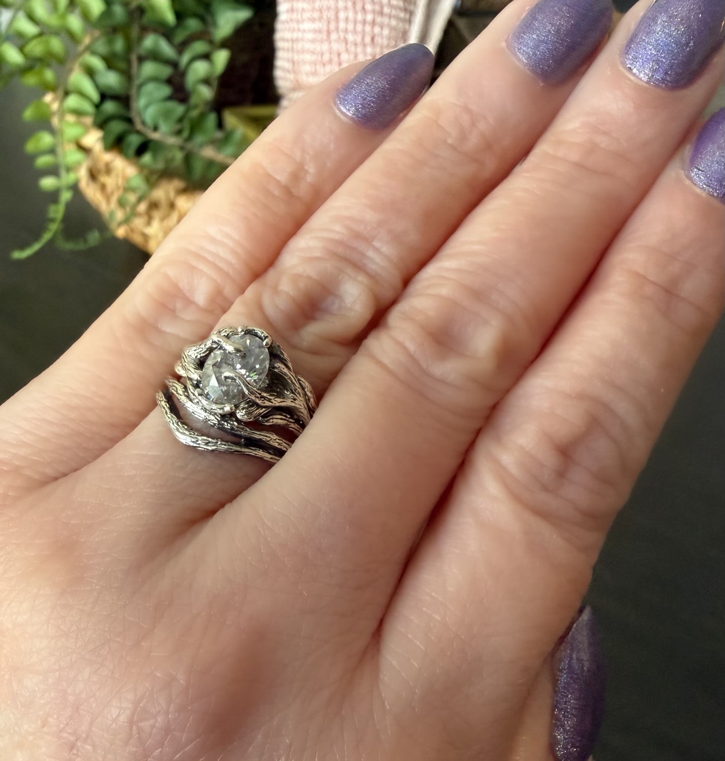 Hand wearing a silver ring with a diamond, against a blurred indoor background