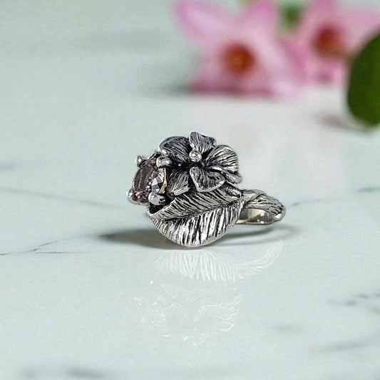 Silver ring with floral design on a marble surface with pink flowers in the background