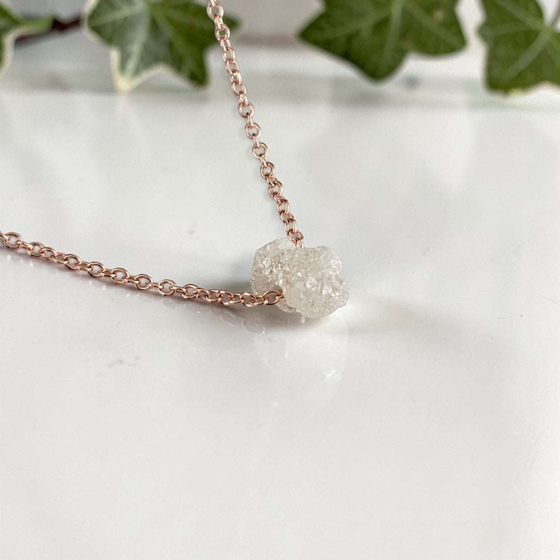 rose gold Necklace with a single raw diamond on a marble surface with ivy leaves in the background