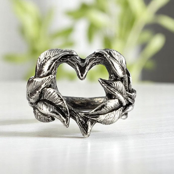 Silver heart-shaped ring with leaf designs on a white surface with a blurred green plant background