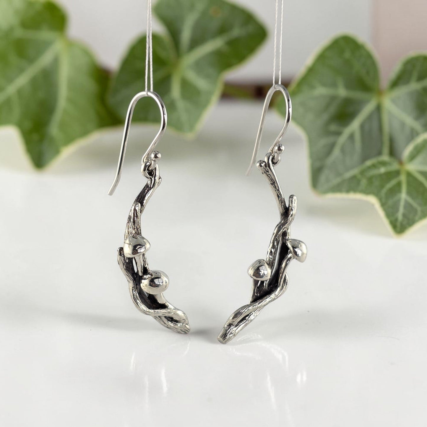Silver branch-shaped earrings with mushrooms on a white surface with green leaves in the background