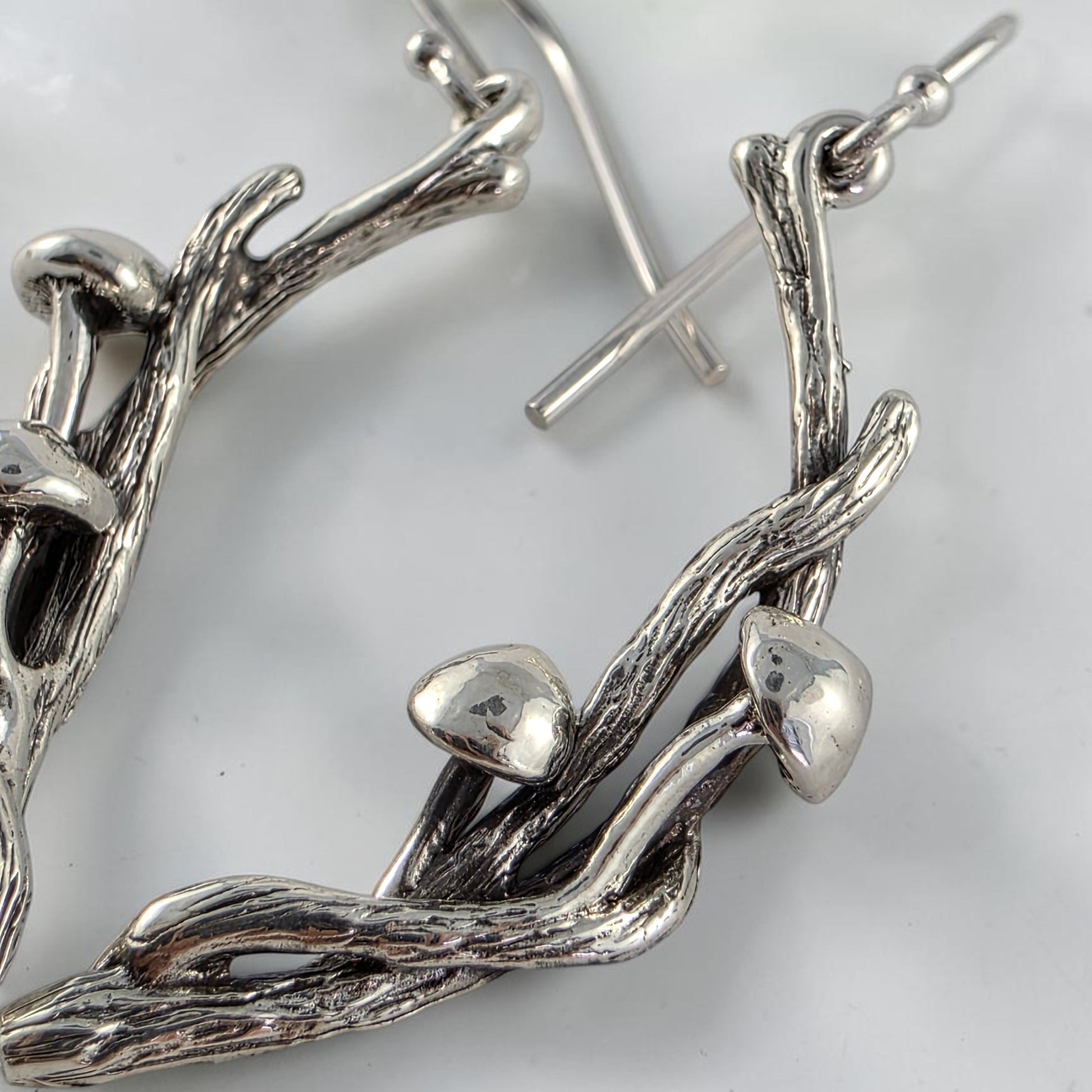 Silver branch-shaped earrings with mushrooms on a white surface with green leaves in the background