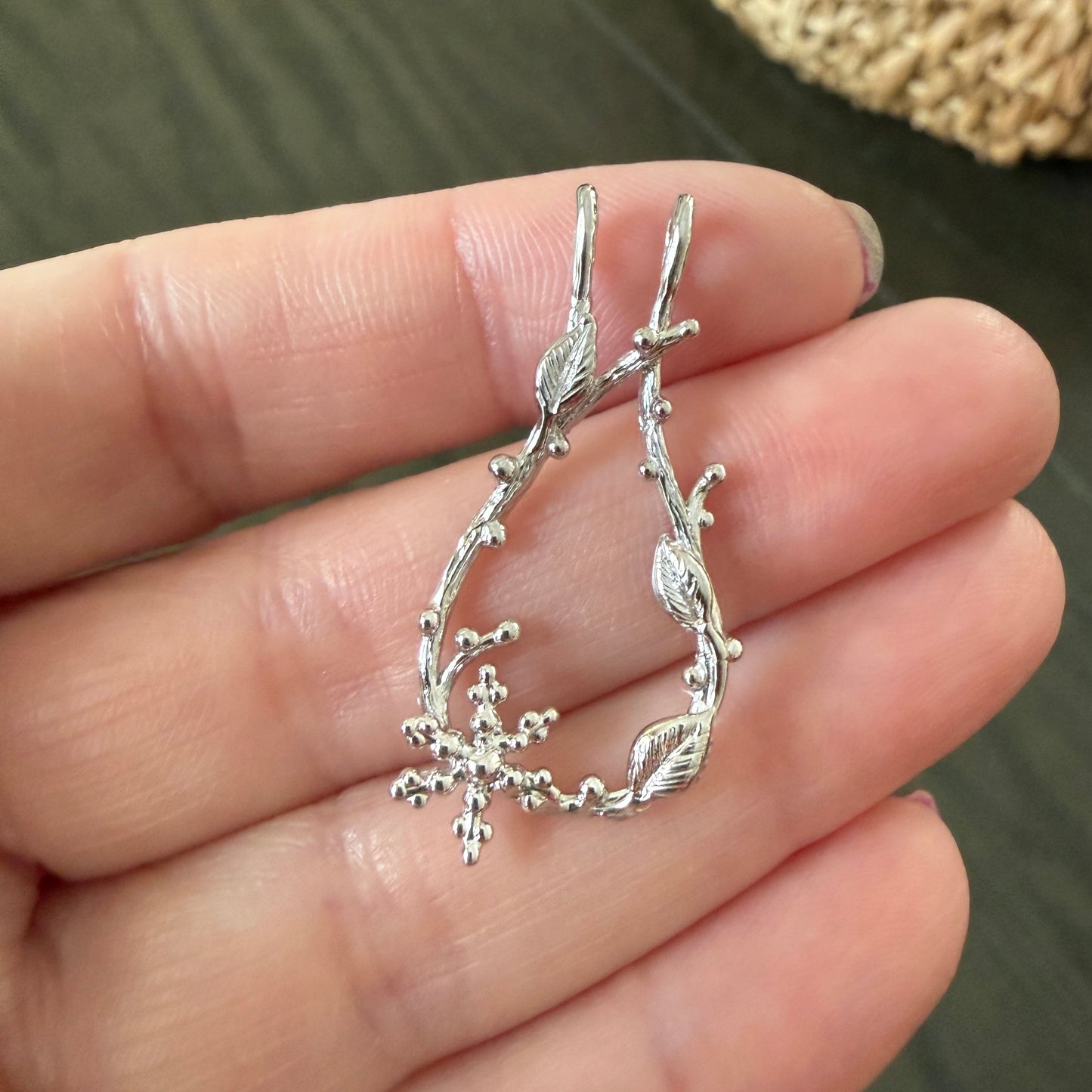 A pair of silver branch-shaped hoop earrings held in a person's hand, with a blurred woven basket and dark tabletop in the background.