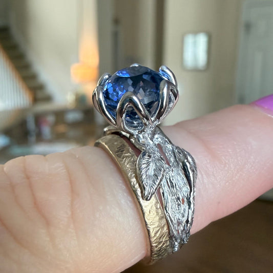 A close-up of a hand wearing a silver ring with a large faceted blue gemstone and textured leaf-like bands, set against a blurred indoor background.