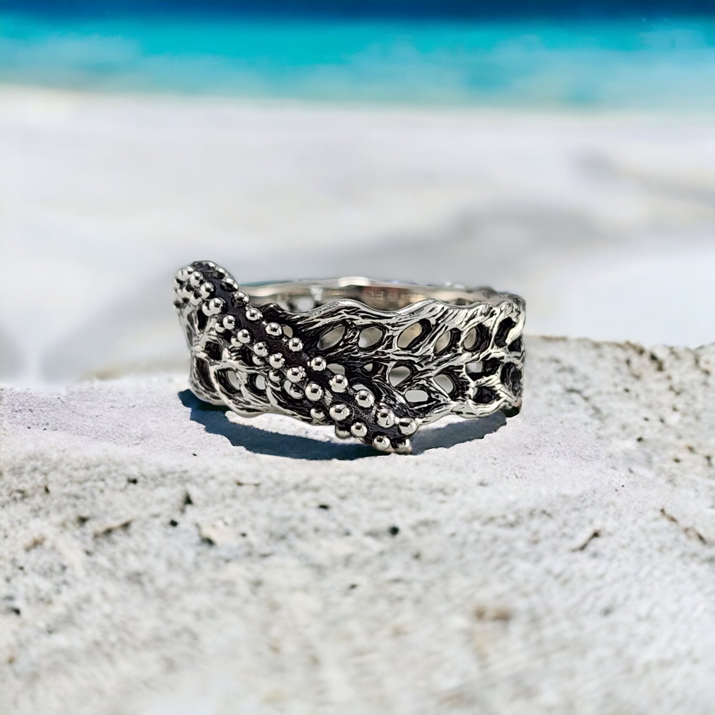 Silver ring with intricate starfish and seafan coral design on a sandy surface with a blurred beach background