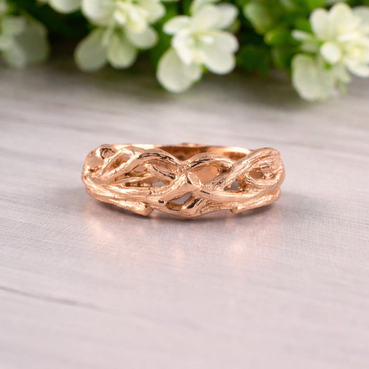 Rose gold ring with braided design on a light surface with flowers in the background