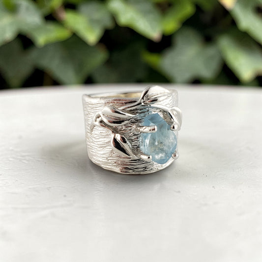 Silver ring with a blue gemstone on a white surface with green foliage in the background