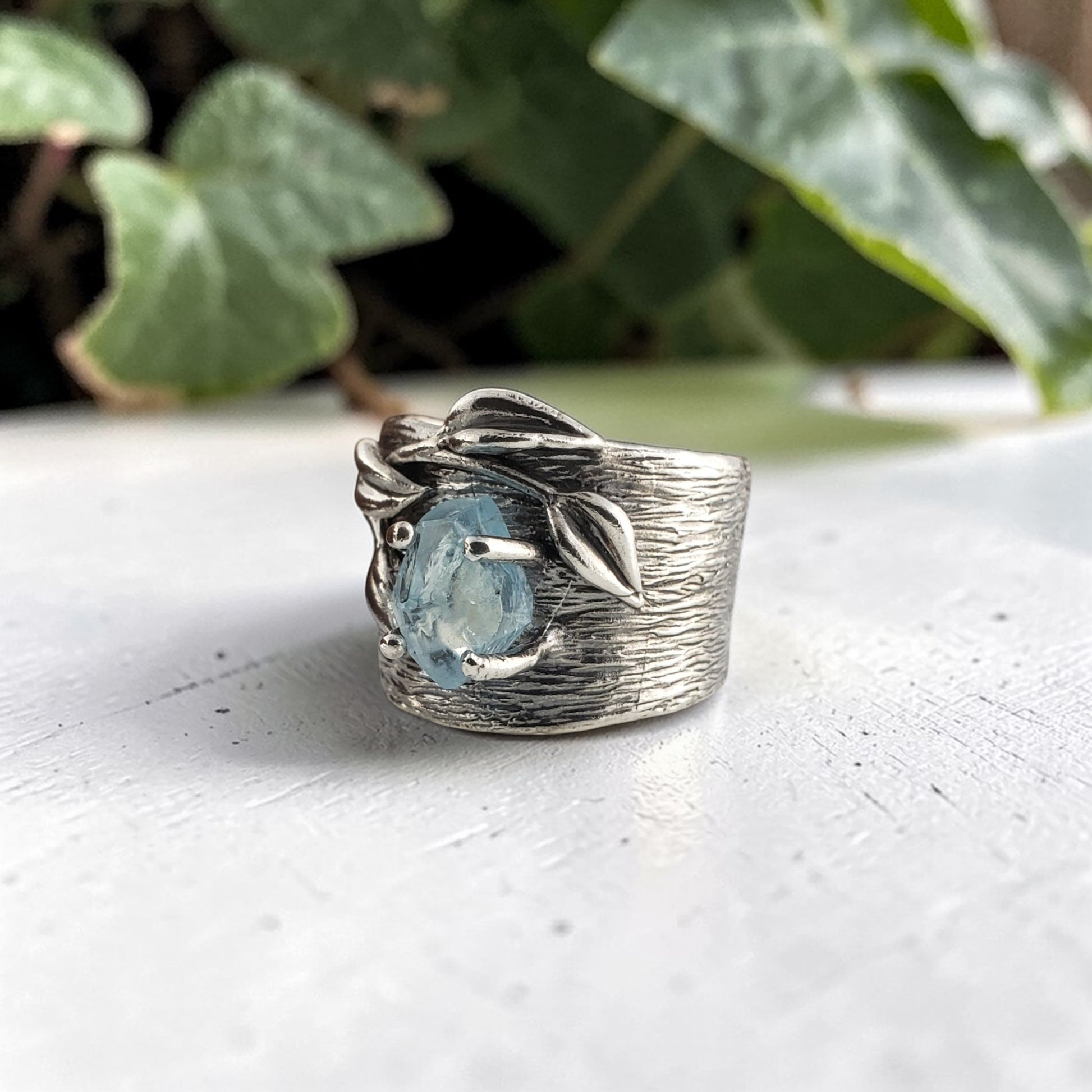 Silver ring with a blue gemstone on a white surface with green leaves in the background