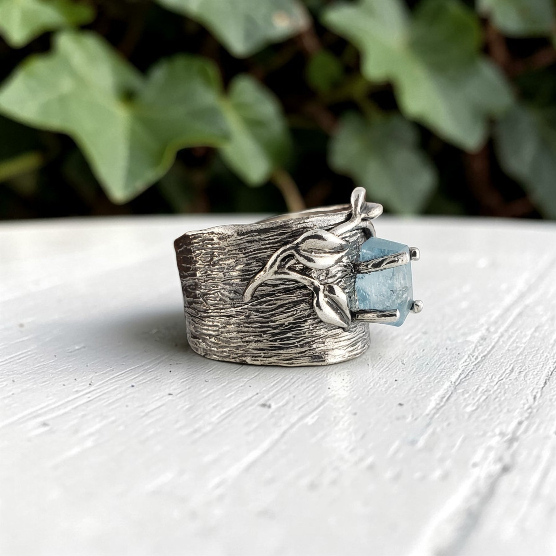Silver ring with a blue gemstone on a white surface with green leaves in the background