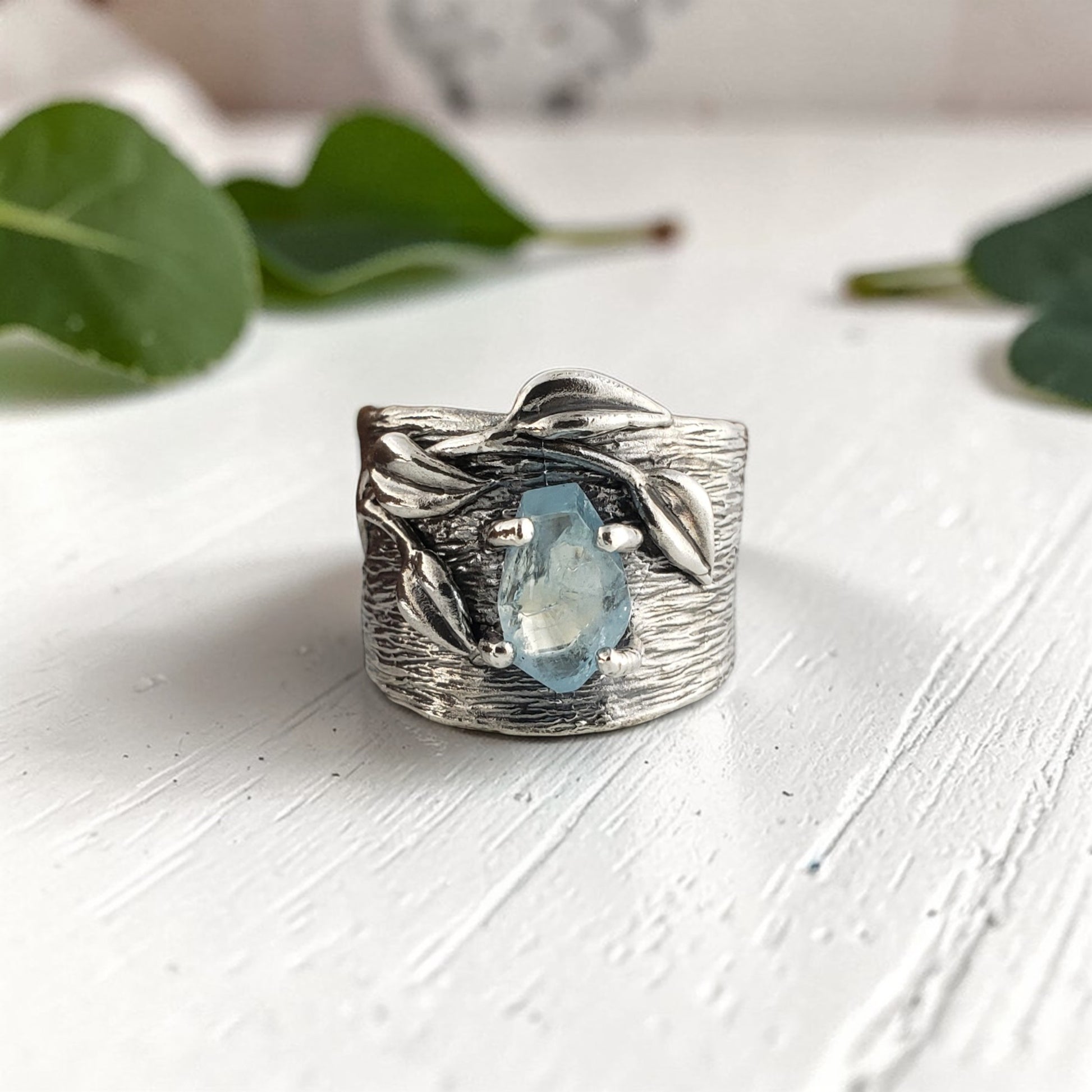 Silver ring with a blue gemstone on a white surface with green leaves in the background